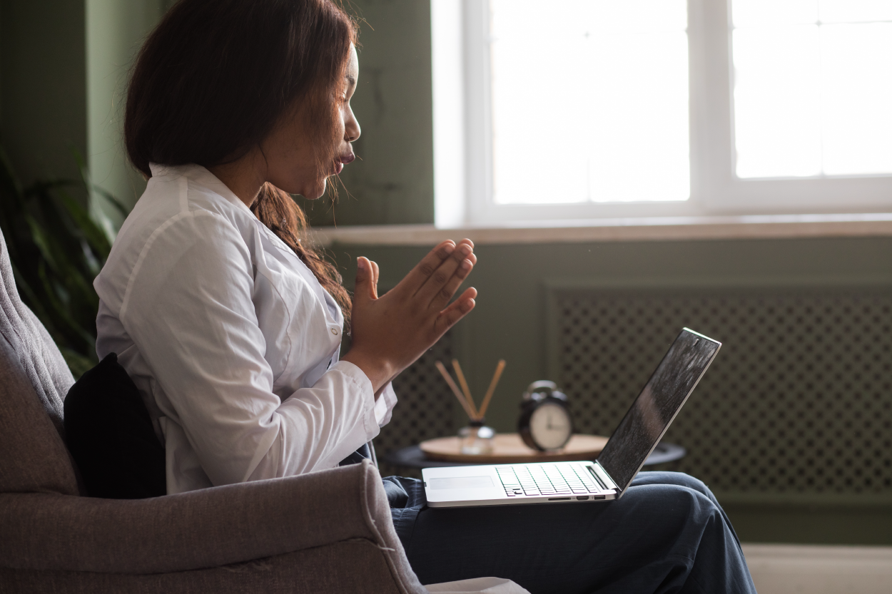 A Black woman sits on a chair with a laptop; her hands are in front of her chest, palms together