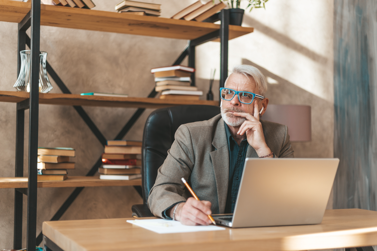 a man wearing glasses sits in front of his laptop and stares off to the side; he holds a pencil over a sheet of paper beside the laptop